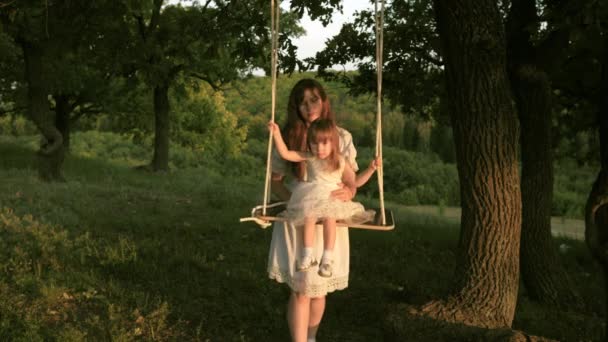 Mutter schüttelt ihre Tochter auf Schaukel unter einem Baum in der Sonne. Nahaufnahme. Mutter und Baby reiten auf einer Seilschaukel auf einem Eichenzweig im Wald. Mädchen lacht, freut sich. Familienspaß im Park, in der Natur. Warmer Sommertag. — Stockvideo