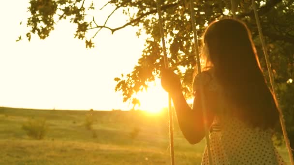 Szczęśliwe dziecko jeździ na huśtawce w parku na dębie w blasku zachodu słońca. Dzieciństwo i doświadczenie w locie. Koncepcja. Gra w locie. Piękna dziewczyna z długimi włosami — Wideo stockowe
