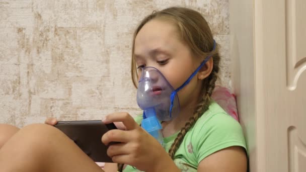 Uma menina doente em uma máscara inala uma garrafa de spray e joga um jogo de computador em um tablet. Um miúdo com uma máscara azul inalou fumos das vias respiratórias para os pulmões. Tratamento da tosse. Close-up — Vídeo de Stock