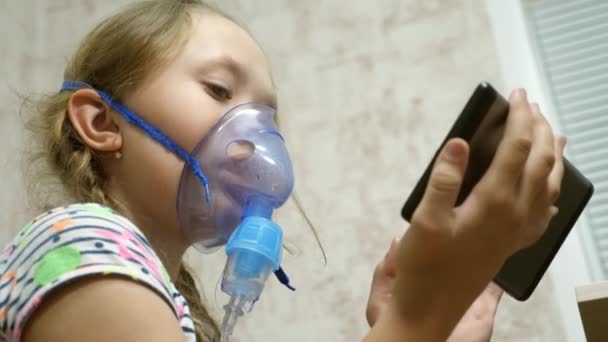 Uma menina doente em uma máscara inala uma garrafa de spray e joga um jogo de computador em um tablet. Um miúdo com uma máscara azul inalou fumos das vias respiratórias para os pulmões. Tratamento da tosse. Close-up — Vídeo de Stock