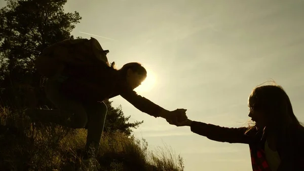 women travelers climb mountain, offer each other a helping hand, helping to climb hill. tourist teamwork. Family of tourists traveling at sunset. sisters with backpacks travel climbing mountain in sun