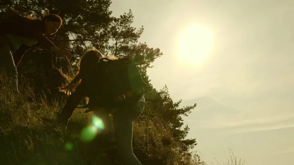 women travelers climb mountain, offer each other a helping hand, helping to climb hill. tourist teamwork. Family of tourists traveling at sunset. sisters with backpacks travel climbing mountain in sun