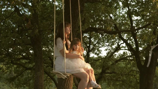 Passeio de mãe e bebê em um balanço de corda em um ramo de carvalho na floresta. Ri menina, alegra-se. Diversão familiar no parque, na natureza. dia quente de verão. Mãe sacode sua filha no balanço sob uma árvore no sol . — Fotografia de Stock