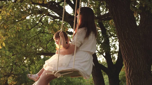 Mor och baby rida på rep Swing på en ek gren i skogen. Flickan skrattar, jublar. Familj kul i parken, i naturen. varma sommardagen. Mamma skakar sin dotter på swing under ett träd i solen. Närbild. — Stockfoto