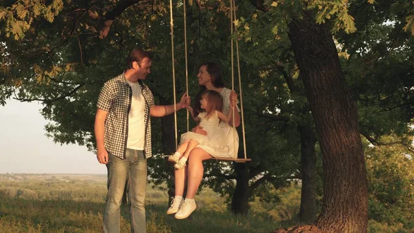 Far skakar mor och barn på ett rep gunga på en ek gren i skogen. Mamma skakar sin dotter när hon svingar sig under ett träd i solen. Pappa skrattar och gläds. Familjekul i parken, i naturen. — Stockfoto