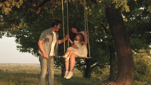 Father shakes mother and child on a rope swing on an oak branch in forest. Mom shakes her daughter on swing under a tree in sun. dad laughs and rejoices. Family fun in park, in nature. — Stock Photo, Image