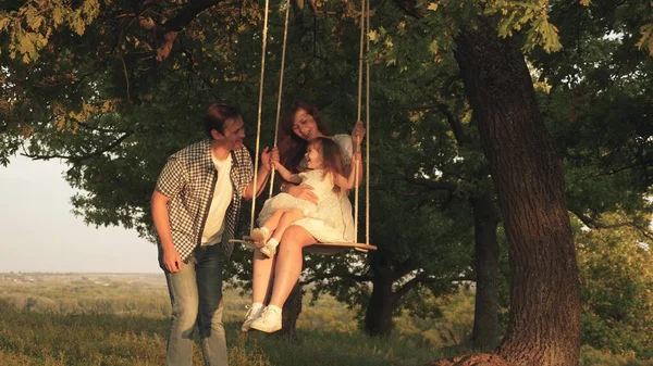 Vater schüttelt Mutter und Kind auf einer Seilschaukel an einem Eichenzweig im Wald. Mutter schüttelt ihre Tochter auf Schaukel unter einem Baum in der Sonne. Papa lacht und freut sich. Familienspaß im Park, in der Natur. — Stockfoto