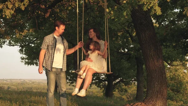 Ο πατέρας κουνάει μητέρα και παιδί σε μια κούνια σε ένα κλαδί βελανιδιάς στο δάσος. Η μαμά κουνάει την κόρη της κάτω από ένα δέντρο στον ήλιο. Ο μπαμπάς γελάει και χαίρεται. Οικογενειακή διασκέδαση στο πάρκο, στη φύση. — Φωτογραφία Αρχείου