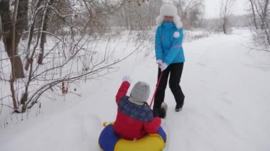 Mutlu anne, beyaz karlı bir yolda kızakla bir çocuğa biniyor. Noel tatilleri. Yetişkinler ve çocuklar için eğlenceli bir oyun. Mutlu aile kavramı. Bir genç Tubing 'e biniyor.