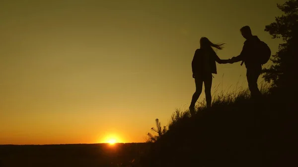 Viajante do sexo masculino segura a mão de uma viajante do sexo feminino ajudando a subir o topo da colina. Os turistas escalam a montanha ao pôr do sol, de mãos dadas. trabalho em equipe de parceiros de negócios. Família feliz em férias . — Fotografia de Stock