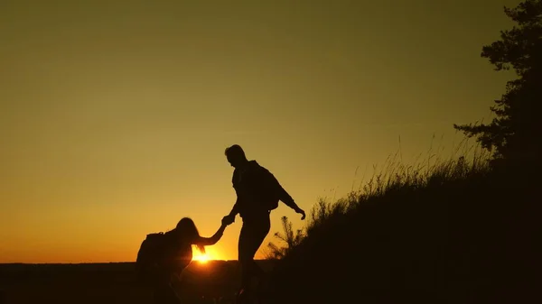 Viajero masculino sostiene la mano de una viajera femenina ayudando a subir la cima de la colina. Los turistas suben a la montaña al atardecer, tomados de la mano. trabajo en equipo de los socios comerciales. Familia feliz de vacaciones . — Foto de Stock