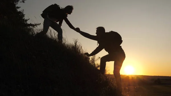 Un viaggiatore maschio tende la mano al viaggiatore femminile che sale su una collina. I turisti scalano la montagna al tramonto, tenendosi per mano. lavoro di squadra di uomini d'affari. Famiglia felice in vacanza . — Foto Stock