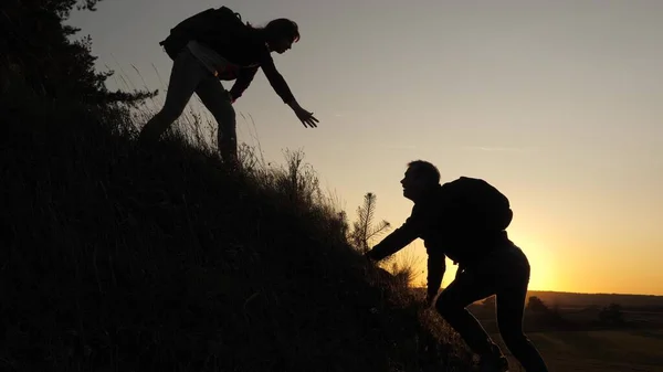 Ένας αρσενικός ταξιδιώτης κρατά το χέρι του σε μια γυναίκα που ανεβαίνει στην κορυφή του λόφου. Οι τουρίστες σκαρφαλώνουν στο βουνό στο ηλιοβασίλεμα, κρατώντας τα χέρια. ομαδικότητα των επιχειρηματικών ανθρώπων. Ευτυχισμένη οικογένεια σε διακοπές. — Φωτογραφία Αρχείου