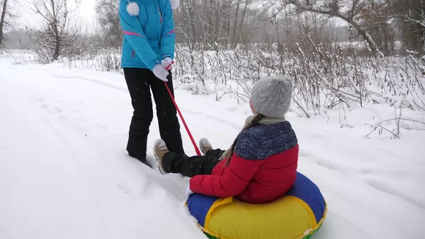 Ευτυχισμένη μητέρα καβαλάει ένα παιδί σε ένα έλκηθρο σε ένα λευκό χιονισμένο δρόμο. Χριστουγεννιάτικες διακοπές. Ένα διασκεδαστικό παιχνίδι για ενήλικες και παιδιά. Η έννοια της ευτυχισμένης οικογένειας. Ένας έφηβος μπαίνει στο Τούμπιγκ. — Φωτογραφία Αρχείου