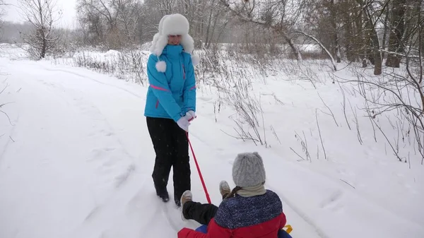 La madre feliz monta a un niño en un trineo en un camino blanco nevado. Vacaciones de Navidad. Un divertido juego para adultos y niños. El concepto de una familia feliz. Un adolescente cabalga en Tubing — Foto de Stock