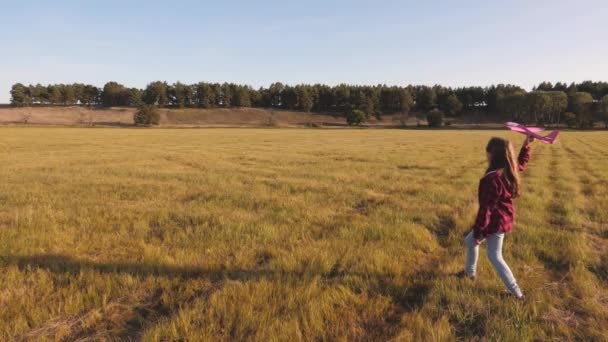 Дівчинка-підліток з довгим волоссям грає на полі з літаком і летить вперед. Дитина мріє стати пілотом. Щаслива дівчина хоче стати пілотом і астронавтом — стокове відео