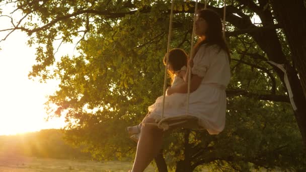 Mom shakes her daughter on swing under a tree in sun. mother and baby ride on a rope swing on an oak branch in forest. Girl laughs, rejoices. Family fun in park, in nature. warm summer day. — Stock Video