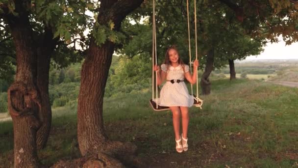 Child swinging on a swing in park in sun. young girl swinging on rope swing on an oak branch. teen girl enjoys flight on swing on summer evening in forest. concept of happy family and childhood. — Stock Video