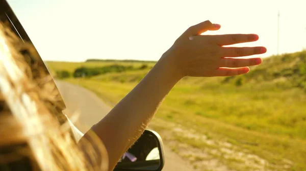 Dziewczyna z długimi włosami siedzi z przodu siedzeniu samochodu, wyciągając rękę z okna i połowu blasku zachodzącego słońca — Zdjęcie stockowe