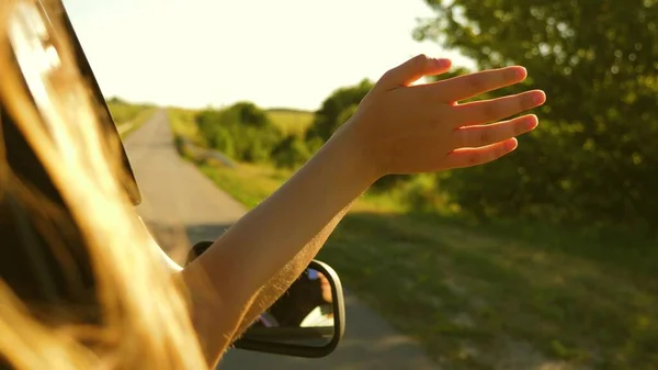 Dziewczyna z długimi włosami siedzi z przodu siedzeniu samochodu, wyciągając rękę z okna i połowu blasku zachodzącego słońca — Zdjęcie stockowe