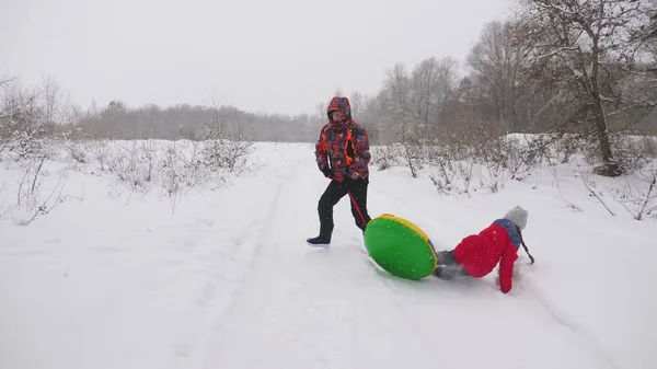 Der glückliche Papa schlittert ein Kind auf einer verschneiten Straße. Weihnachtsferien. Der Vater spielt mit seiner Tochter im Winterpark. das Konzept einer glücklichen Familie. Teenager fährt in Schläuchen — Stockfoto