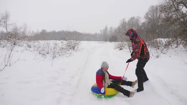 Gelukkige vader sleeën een kind op een besneeuwde weg. Kerstvakantie. Vader speelt met zijn dochter in een winterpark. Het concept van een gelukkig gezin. Een tiener rijdt in Tubing — Stockfoto