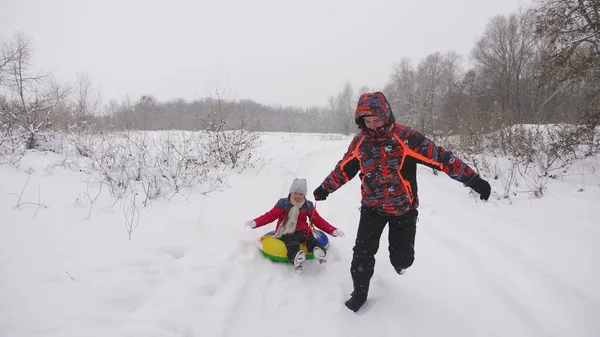 Ο χαρούμενος μπαμπάς σέρνει ένα παιδί σε ένα χιονισμένο δρόμο. Χριστουγεννιάτικες διακοπές. Ο πατέρας παίζει με την κόρη του σε ένα χειμερινό πάρκο. Η έννοια της ευτυχισμένης οικογένειας. Ένας έφηβος μπαίνει στο Τούμπιγκ. — Φωτογραφία Αρχείου