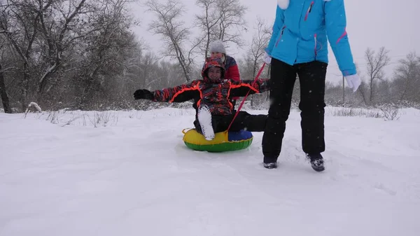 Mamma och dotter åker pulka pappa i vinterskogen, i snöfall. Glada föräldrar och barn leker i julparken. Familjen leker i vinterparken på julhelgen. — Stockfoto