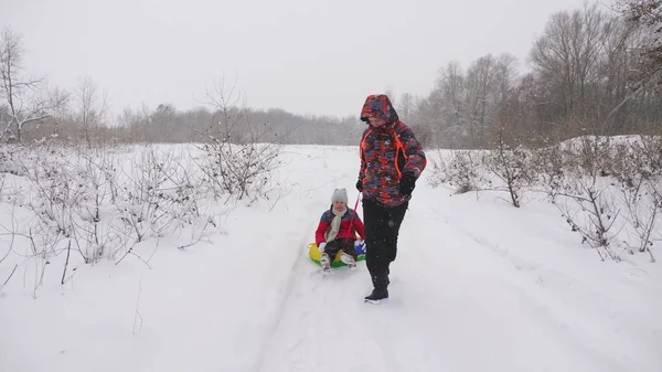 Mutlu baba, karlı bir yolda bir çocuğu kızaklıyor. Noel tatilleri. Baba, kızıyla kış parkında oynuyor. Mutlu aile kavramı. Bir genç Tubing 'e biniyor. — Stok fotoğraf