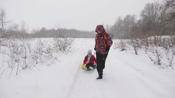 Der glückliche Papa schlittert ein Kind auf einer verschneiten Straße. Weihnachtsferien. Der Vater spielt mit seiner Tochter im Winterpark. das Konzept einer glücklichen Familie. Teenager fährt in Schläuchen — Stockfoto