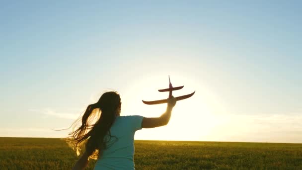 幸せな女の子は日没の光の中でフィールド上のおもちゃの飛行機で実行されます。子供はおもちゃの飛行機で遊ぶ。10代の頃はパイロットになることを夢見ていました。少女はパイロットと宇宙飛行士になりたがっている。スローモーション — ストック動画