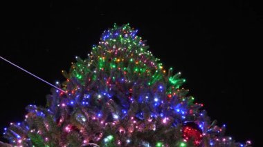 Noel tatili konsepti. Çelenklerle süslenmiş güzel, uzun bir Noel ağacı ve gece göğüne karşı çok renkli toplar. Biraz kar yağar. Çelenklerle süslenmiş Noel ağacı.