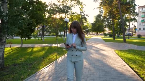 Mujer en traje de luz va a trabajar con la tableta en la mano. Hermosa mujer de negocios camina por el callejón en el parque y comprueba el correo electrónico en la tableta . — Vídeo de stock