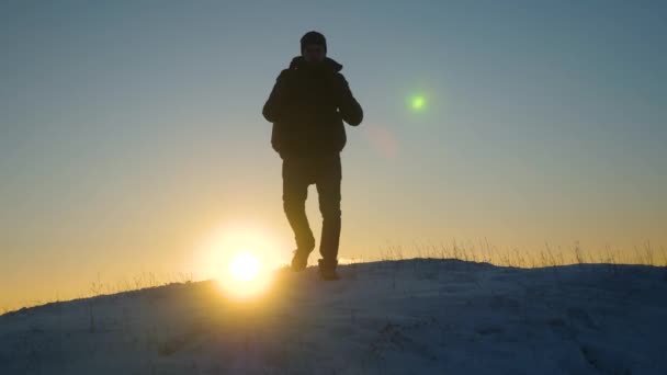 Alpinista viaja senderismo. Un viajero desciende en una montaña nevada desde la cima, a la luz del atardecer. turista va a la victoria superando las dificultades. viajero va por la ladera nevada a una colina nevada en el sol . — Vídeo de stock