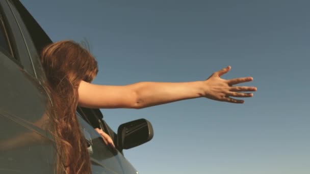 Happy female traveler rides car along a country road. hand from car window catches the wind. Girl with long hair is sitting in front seat of car, stretching her arm out window and catching glare sun — Stock Video