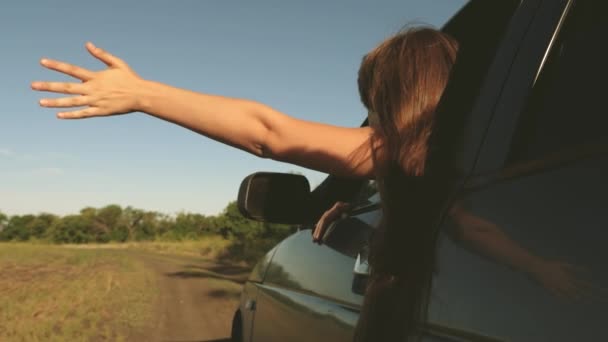 Happy female traveler rides car along a country road. hand from car window catches the wind. Girl with long hair is sitting in front seat of car, stretching her arm out window and catching glare sun — Stock Video