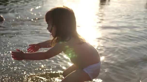 Spruzzi di bambino in acqua. spruzzando acqua. Bambino carino gioca felicemente sulla spiaggia. bambino felice si bagna al tramonto su un fiume, mare, lago. concetto di infanzia felice . — Video Stock