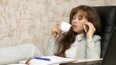 ofis iş yerinde kahve fincan ile akıllı telefonda konuşan kız. bilgisayar çalışması. güzel iş kadın masa üzerinde çıplak ayaklar ile bir sandalyede oturan rahatlatıcı ve çay içme, kahve.