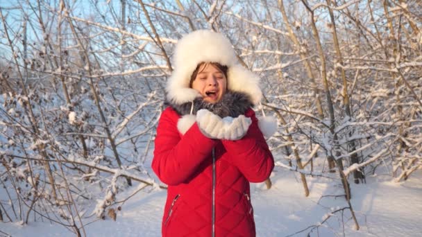Щаслива концепція дитинства. дівчина дме сніжинки на заході сонця і посміхається в зимовому парку. Повільний рух. молода дівчина дме сніжинки з її рук, в лісі. дитина грає взимку на різдвяні канікули . — стокове відео