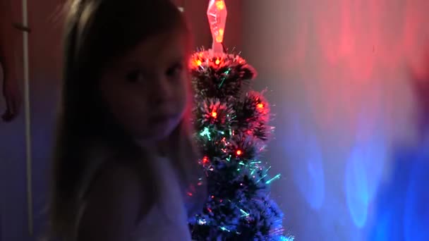 Pequena criança joga pela árvore de Natal no quarto das crianças. filha examina guirlanda na árvore de Natal. bela árvore de Natal artificial. conceito de infância feliz. Família joga para feriados de Natal — Vídeo de Stock
