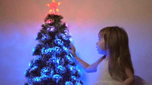 Маленький ребенок играет у елки в детской комнате. дочь осматривает гирлянду на елке. красивая искусственная елка. счастливая детская концепция. Семейные спектакли на рождественские праздники — стоковое видео