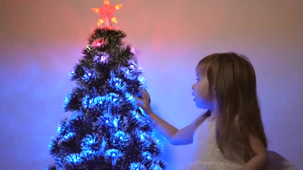 Pequena criança joga pela árvore de Natal no quarto das crianças. filha examina guirlanda na árvore de Natal. bela árvore de Natal artificial. conceito de infância feliz. Família joga para feriados de Natal — Vídeo de Stock