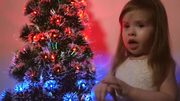 A lánya virágfüzért vizsgál a karácsonyfán. kisgyerek játszik karácsonyfa a gyermek szobában. Gyönyörű mesterséges karácsonyfa. Boldog gyermekkori koncepciót. Családi játékok a karácsonyi szünetben — Stock videók