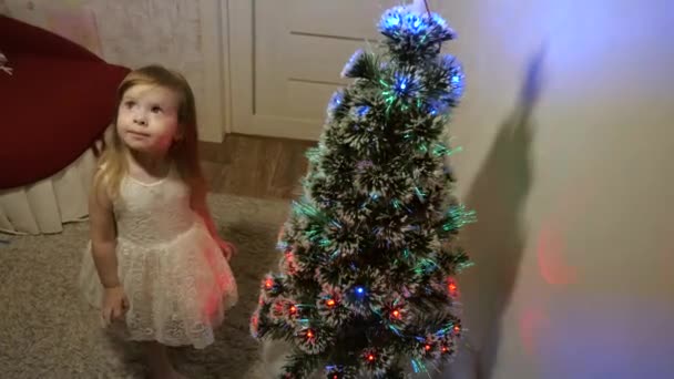 Concetto di infanzia felice. bambino piccolo gioca vicino all'albero di Natale nella stanza dei bambini. bellissimo albero di Natale artificiale. figlia esamina ghirlanda sull'albero di Natale. Famiglia gioca per le vacanze di Natale — Video Stock