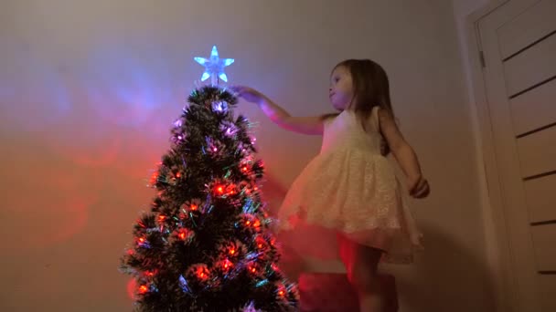 Kind onderzoekt een kerstster op een kerstboom. klein meisje speelt in de buurt van een kerstboom in een kinderkamer. mooie kunstmatige kerstboom. Het concept van een gelukkige jeugd. — Stockvideo