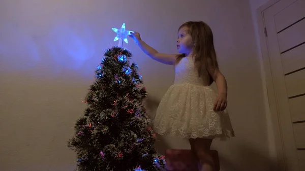 Enfant examine une étoile de Noël sur un arbre de vacances. petite fille joue près d'un arbre de Noël dans une chambre d'enfant. magnifique arbre de Noël artificiel. Le concept d'une enfance heureuse . — Photo