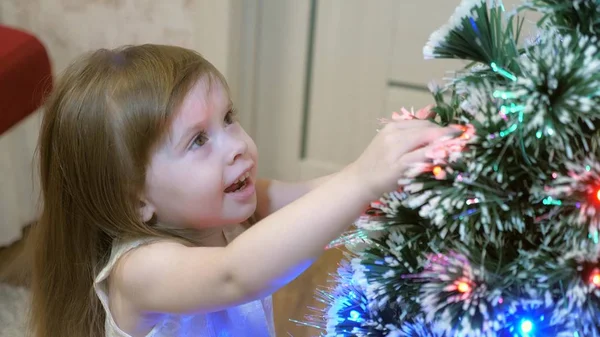小女孩在圣诞树上检查一个花环。 孩子们在孩子们房间里的圣诞树旁玩耍。 美丽的人造圣诞树。 快乐童年的概念。 全家都在过圣诞节 — 图库照片