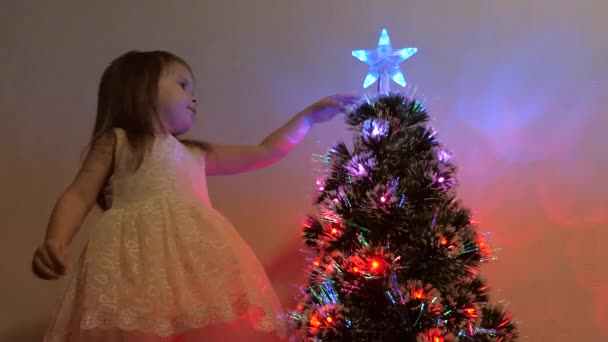 Niño examina una estrella de Navidad en un árbol de vacaciones. La niña juega cerca de un árbol de Navidad en una habitación para niños. hermoso árbol de Navidad artificial. El concepto de una infancia feliz . — Vídeos de Stock