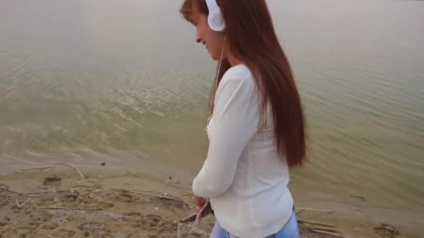 Chica joven camina en la playa con auriculares y escucha música en la tableta mientras baila y se ríe con una sonrisa abierta — Vídeos de Stock