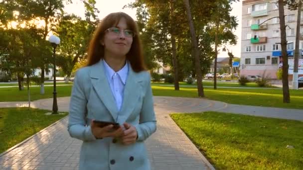 Chica en traje de luz va a trabajar con la tableta en la mano. Hermosa mujer de negocios camina por el callejón en el parque y comprueba el correo electrónico en la tableta . — Vídeos de Stock